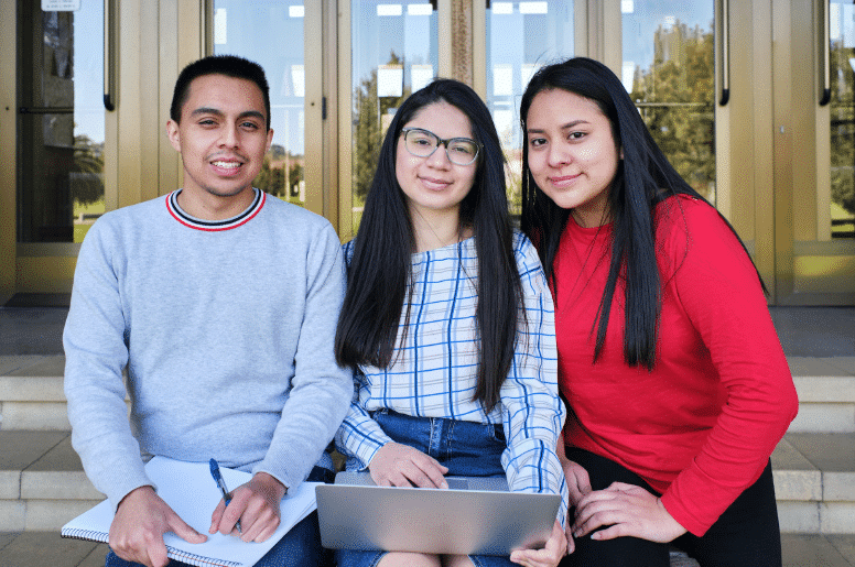 three students sitting on steps and smiling with the middle student on the computer